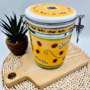 May include: A yellow ceramic canister with a metal clamp lid, featuring brown coffee bean illustrations and the text "Java" and "Mocha" in black script. The lid has a central coffee bean graphic surrounded by a decorative leaf border pattern.