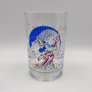 May include: A clear glass tumbler featuring a blue, red, and yellow graphic of Mickey Mouse in a wizard robe pointing towards a large sphere. The design includes fireworks and the text Remember the Magic etched into the base of the glass.