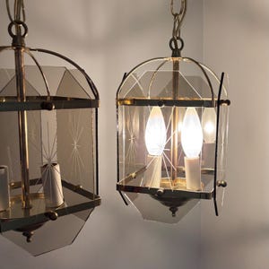May include: Two vintage pendant lights with a brass finish and geometric glass panels. The glass has a starburst design. Each light fixture has a chain for hanging and contains multiple light bulbs.