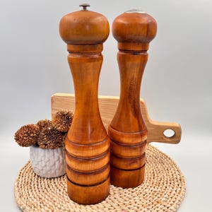 May include: A pair of wooden salt and pepper mills in a warm brown hue. The mills feature a traditional design with a rounded top and a slender, grooved body. They are presented with a small white vase, dried flowers, a wooden chopping board, and a woven mat.