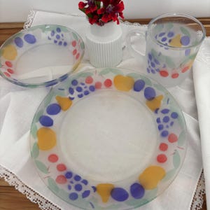 Puede incluir: Un juego de vajilla de vidrio transparente con un diseño de frutas. El juego incluye un cuenco, un plato y una taza. El diseño de frutas presenta formas amarillas, azules y rojas con hojas verdes. El juego está sobre un paño blanco.