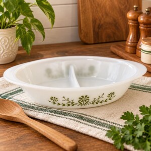 Peut inclure: Un plat de service ovale en verre opalin blanc divisé en deux sections par une paroi centrale. L'extérieur est orné d'un motif floral vert représentant des marguerites et des tiges feuillues, avec des poignées intégrées sur les côtés.