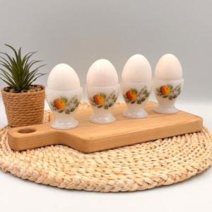 May include: Four white milk glass egg cups, each featuring a painted orange peach and green leaves on the exterior. The egg cups are arranged in a row on a light-colored wooden cutting board, with each cup holding a single white egg.