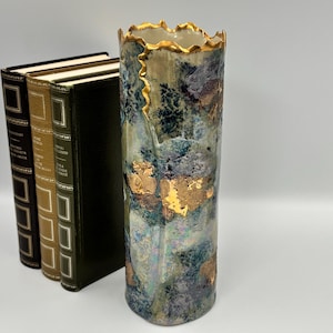 Peut inclure: Un vase cylindrique en céramique avec une surface texturée et marbrée dans des tons de bleu, vert et gris, orné d'accents de feuilles d'or irréguliers et d'un bord supérieur dentelé doré. Le vase est posé à côté de trois livres reliés.