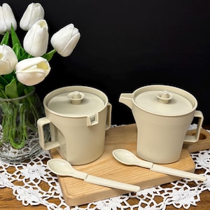 May include: Two beige serving pitchers with lids and handles, accompanied by two matching spoons, are arranged on a wooden cutting board. The pitchers and spoons are made of a light-colored material. A vase of white tulips is in the background.