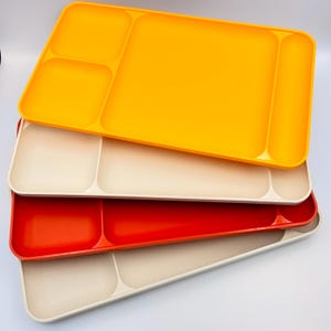 May include: A stack of colorful plastic trays in shades of yellow, cream, red, and beige. Each tray has a rectangular shape with multiple compartments, ideal for serving food or organizing items. The trays are stacked on top of each other.