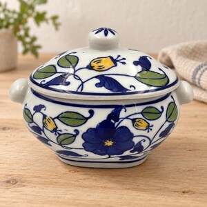 May include: A white ceramic lidded bowl featuring a hand-painted floral pattern in deep blue, sage green, and yellow. The bowl has two small side handles and a matching lid with a central knob, decorated with vines, leaves, and large blossoms.