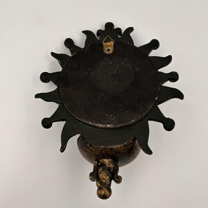 May include: A black sun-shaped wall plaque with a circular center and radiating points. The back features a metallic triangular hanging bracket attached to the top. The lower portion has a textured, mottled gold and black finish.