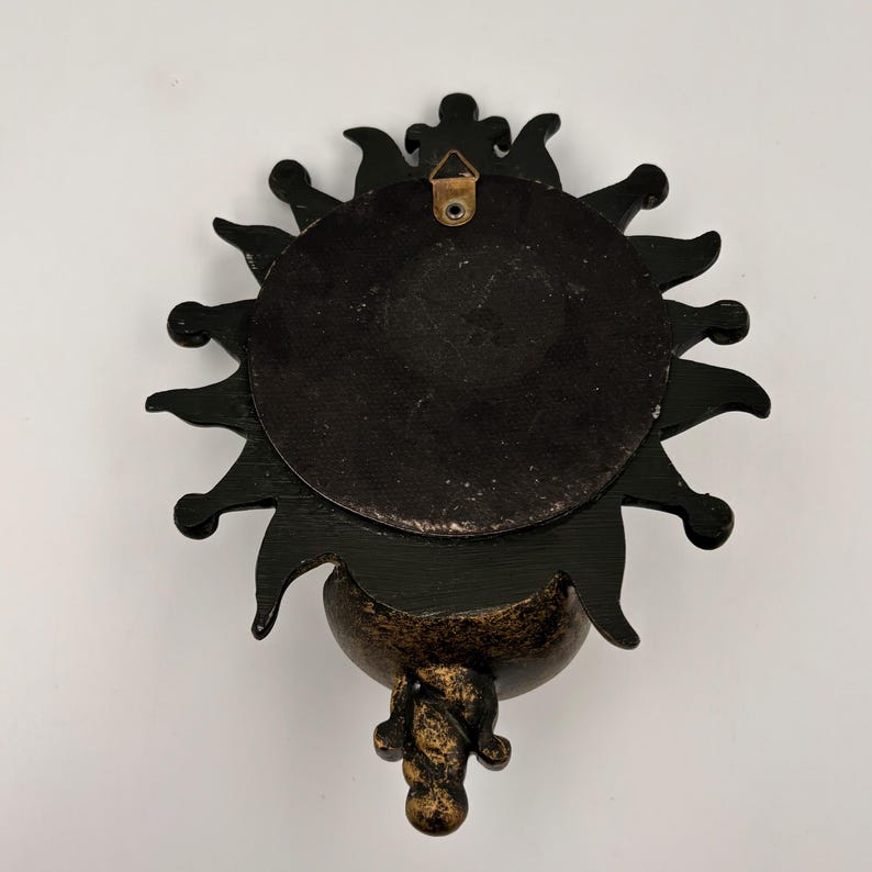May include: A black sun-shaped wall plaque with a circular center and radiating points. The back features a metallic triangular hanging bracket attached to the top. The lower portion has a textured, mottled gold and black finish.