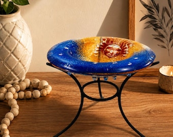 Könnte beinhalten: Eine kreisförmige Glasschale mit einem Farbverlauf von tiefem Blau zu warmem Bernstein und einem geformten Sonnengesicht-Design in der Mitte. Die Schale ruht auf einem schwarzen Metallständer mit vier geschwungenen Beinen und einem Ringfuß.