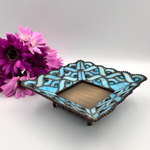 Peut inclure: Un plateau décoratif carré avec un motif en vitrail bleu clair et un intérieur de couleur bronze. Le plateau a une bordure détaillée et ornée avec de petites perles foncées. Un bouquet de fleurs violettes est en arrière-plan.