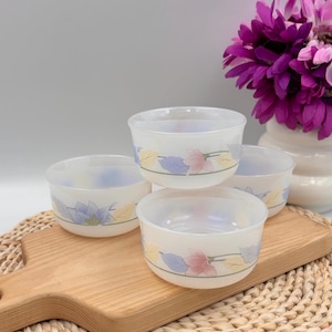 May include: A set of three small, round, white milk glass bowls featuring a delicate floral pattern with pastel pink, blue, and yellow leaves and petals encircling the exterior. The bowls have a smooth, translucent finish and a simple rim.