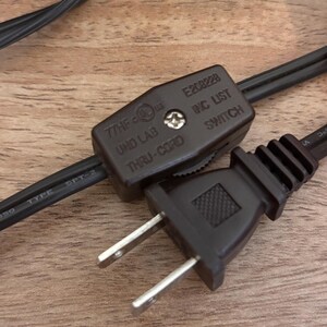 K&ouml;nnte beinhalten: Ein dunkelbraunes Stromkabel mit einem integrierten Daumenradschalter und einem zweipoligen Stecker. Das Schaltergeh&auml;use ist mit dem Text 77HF, E208228, UND LAB, INC LIST und THRU-CORD gepr&auml;gt. Das Kabel ist als Typ SPT-2 f&uuml;r die Stromverbindung gekennzeichnet.