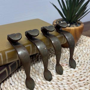 以下が含まれることがあります： 4つのアンティークブロンズトーンの金属製プル。各プルは、ハート型のトップと涙型のベースを備えた湾曲したハンドルを備えています。プルは一列に配置され、本の上に置かれています。