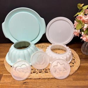 Puede incluir: Una colección de utensilios de cocina vintage, que incluye una fuente para servir azul claro con asas, dos tapas blancas y dos moldes decorativos. El juego también incluye un molde acanalado azul claro y tres tapas decorativas pequeñas y transparentes con diseños en relieve.