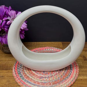 May include: A white ceramic vase with a circular handle. The vase has a shallow, oval-shaped base and a smooth, glossy finish. The vase is sitting on a colorful, woven placemat. Purple flowers are visible in the background.