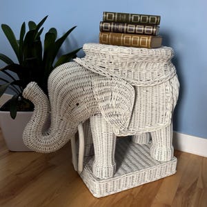 May include: A white wicker elephant-shaped side table with a shelf and a flat top. Three books with gold and green covers are stacked on top. The table is placed on a wooden floor, with a potted plant in the background.