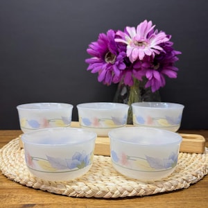 Pode incluir: Cinco tigelas de vidro fosco com desenhos florais em rosa, azul e amarelo. As tigelas estão dispostas sobre um descanso de mesa tecido e uma bandeja de madeira, com um vaso de flores roxas ao fundo.