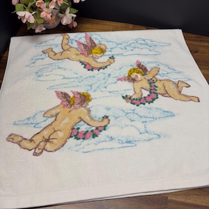 May include: White bath towel featuring three cherubs with pink wings and floral garlands, floating among blue clouds. The cherubs are depicted in a light skin tone, adding a touch of whimsy to the design.