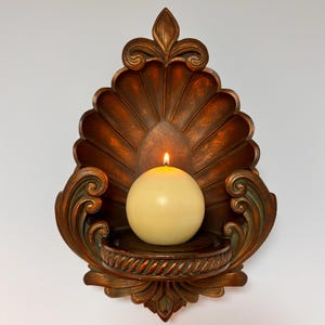 May include: A wall-mounted candle sconce featuring a bronze-toned, shell-shaped backplate with ornate scrollwork and a rope-textured circular base. A single, round, cream-colored sphere candle sits in the center, emitting a small, bright flame.