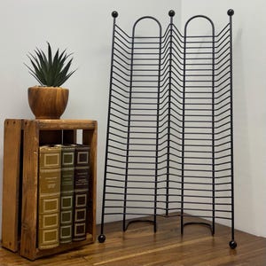 Puede incluir: Un soporte de discos de metal negro con un diseño decorativo. El soporte está sobre una superficie de madera junto a una caja de madera con libros y una planta en maceta.