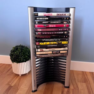 May include: A silver and black vertical media storage rack with a curved frame, featuring the brand name Atlantic printed on the base. The rack holds a stack of various DVD cases with visible titles including The Usual Suspects, Speed, and Rain Man.