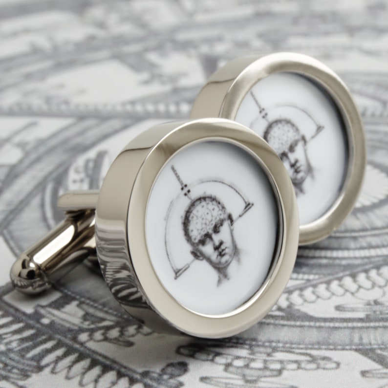 May include: A pair of silver cufflinks with a circular design. Each cufflink features a black and white illustration of a human head with a diagram of the brain. The cufflinks have a polished, reflective finish.