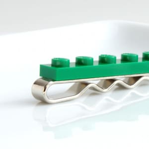 May include: A green Lego brick tie clip with a silver-coloured metal clip. The tie clip features a rectangular green Lego brick with six circular studs. The metal clip has a wavy design.