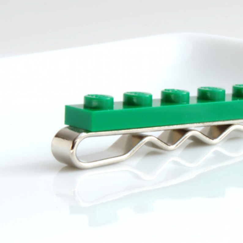 May include: A green Lego brick tie clip with a silver-coloured metal clip. The tie clip features a rectangular green Lego brick with six circular studs. The metal clip has a wavy design.