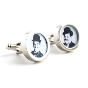 May include: A pair of silver cufflinks with a round, white background and a black and white image of Charlie Chaplin in a black hat.