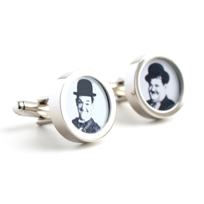 May include: A pair of silver cufflinks with a round, white background and a black and white image of Charlie Chaplin in a black hat.
