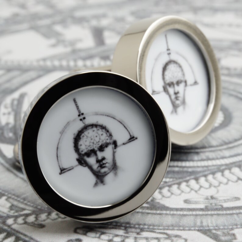 May include: A pair of silver cufflinks with a black and white illustration of a human head with a brain diagram. The cufflinks have a circular design with a polished silver finish and a black border.