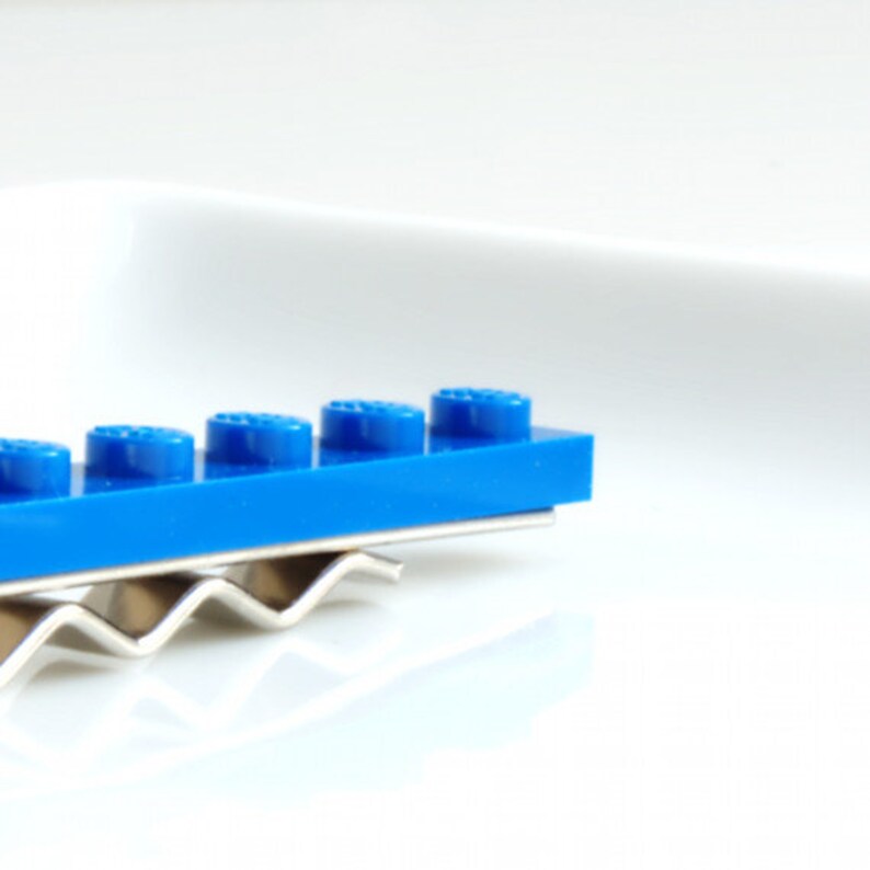 May include: A blue Lego tie clip with a silver metal clip. The Lego piece is made up of six individual bricks.