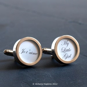 May include: A pair of silver cufflinks with a white background and black text. One cufflink says "Je t'aime" and the other says "My Little Girl".