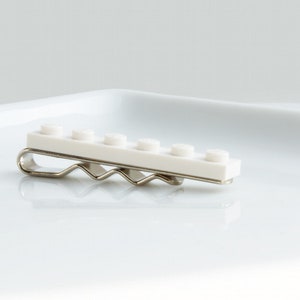 May include: A white tie clip made from six connected Lego bricks. The clip is attached to a silver metal bar.