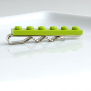 May include: A lime green Lego brick tie clip with a silver metal clip. The tie clip features a rectangular Lego brick with five circular studs. The metal clip has a wavy design.
