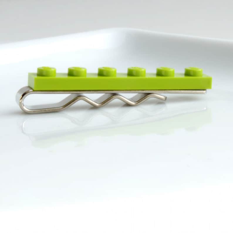 May include: A lime green Lego brick tie clip with a silver metal clip. The tie clip features a rectangular Lego brick with five circular studs. The metal clip has a wavy design.