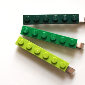 May include: Three green and lime green Lego brick hair clips. The clips are made of plastic and have a metal clasp. The Lego bricks are arranged in a row and have the Lego logo on each brick.