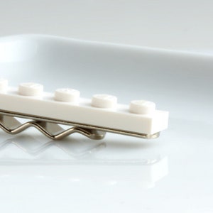 May include: A white hair clip with a row of white, circular studs, similar to toy building blocks. The clip is attached to a silver-coloured metal base, creating a playful and unique accessory.