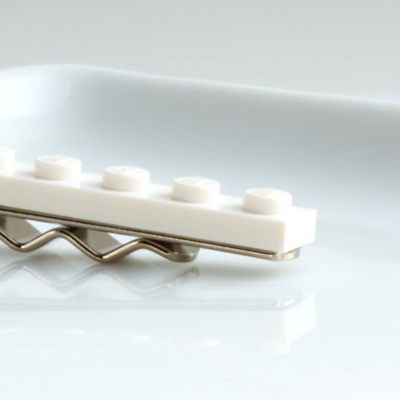 May include: A white hair clip with a row of white, circular studs, similar to toy building blocks. The clip is attached to a silver-coloured metal base, creating a playful and unique accessory.
