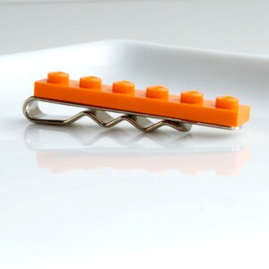 May include: An orange Lego brick hair clip with a silver-coloured metal clip. The clip has a row of six raised studs. This playful hair accessory is a unique item.