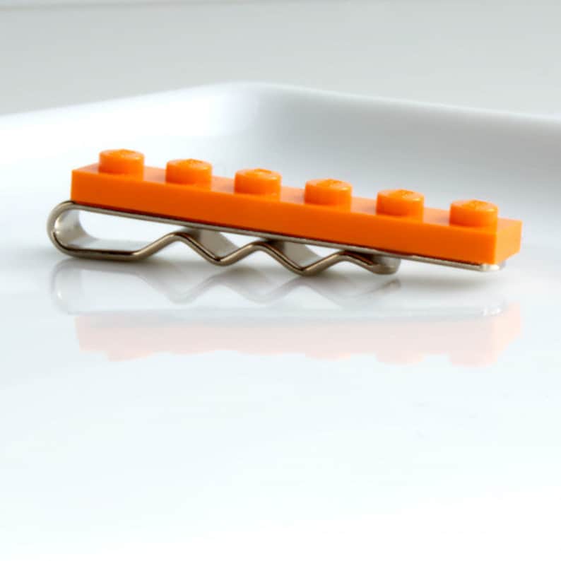 May include: An orange Lego brick hair clip with a silver-coloured metal clip. The clip has a row of six raised studs. This playful hair accessory is a unique item.