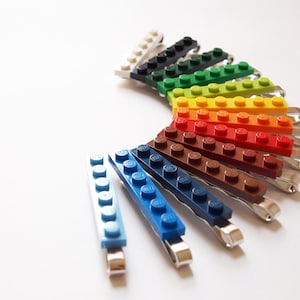 May include: A selection of hair clips, each with a Lego brick design in a range of colours. The clips are arranged in a semi-circle, displaying a spectrum of hues including white, black, green, yellow, and blue. The clips are made of metal.