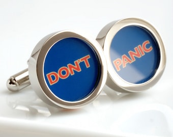 May include: A pair of silver cufflinks with a blue circular face. The text "DON'T PANIC" is written in red letters on the blue background. The cufflinks are on a white surface.