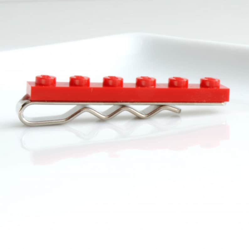 May include: A red Lego brick tie clip with a silver metal clasp. The clip features a classic Lego brick design with six raised studs. A unique accessory for those who enjoy building toys. The clip is approximately 5 cm long.