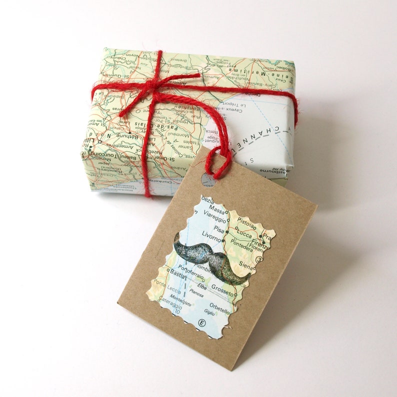 May include: A gift wrapped in map-printed paper, tied with red twine. A brown tag with a map-shaped cutout and a mustache illustration is attached. A creative and unique gift presentation.