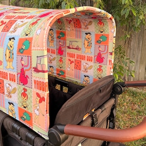 May include: A stroller canopy featuring a colorful patchwork design with cartoon characters and the phrase "Yabba Dabba Doo." The canopy is attached to a brown stroller with a leather-look handle.
