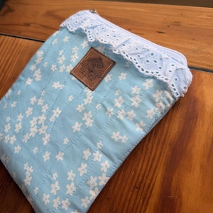 May include: A light blue quilted pouch featuring a white floral pattern, a white eyelet lace trim along the top zipper, and a square brown leather-like patch centered on the front with an illustration of an open book and the text Just One More Chapter.