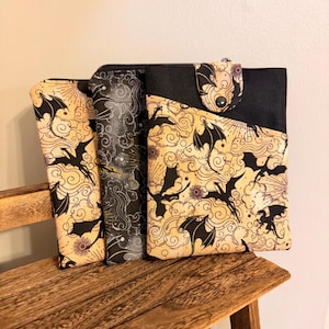 May include: Three fabric pouches featuring a black dragon and cloud pattern on a tan background, with one pouch displaying a black and grey swirling design. Each pouch has a black zipper closure and a snap button flap on the front.