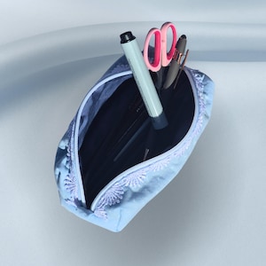 Könnte beinhalten: Ein hellblaues Federmäppchen mit floraler Spitzenborte, geöffnet, um Stifte, Bleistifte und eine Schere zu zeigen. Ein schwarzer Marker und eine rosa Schere sind sichtbar. Das Etui befindet sich auf einem hellblauen Hintergrund.